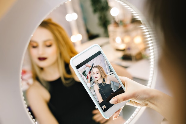 Elegant pretty young woman making photo on phone in mirror during making hairstyle in hairdresser salon. stylish fashionable model, preparing to party, celebration, luxury look