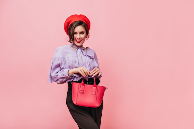 Flirtatious woman in beret, blouse and trousers holding red bag on pink background.